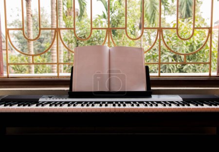 Electric piano with empty sheet music near the big window overlooking green garden, front view