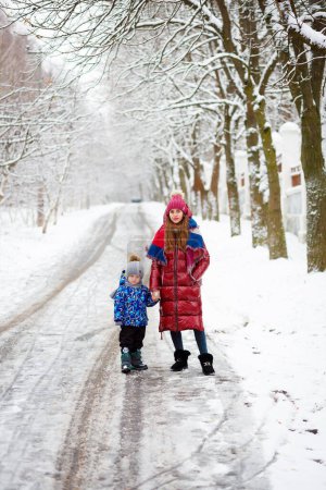 Foto de Hermosa madre joven vestida con chaqueta roja con un niño en invierno cerca de la casa con un árbol de Navidad cubierto de nieve - Imagen libre de derechos