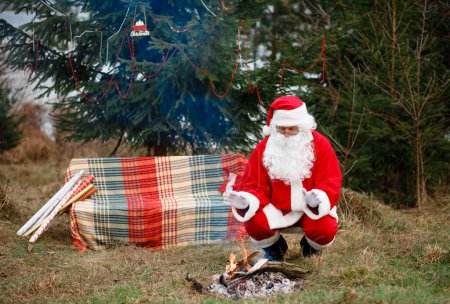 Foto de Santa Claus sits in nature by the fire near the Christmas trees and warms his hands. Red suit with a white beard. Winter holidays in the world - Imagen libre de derechos