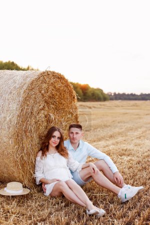 Foto de A pregnant woman with her husband on a summer walk in a wheat field. Fashionable summer clothes. White dress - Imagen libre de derechos