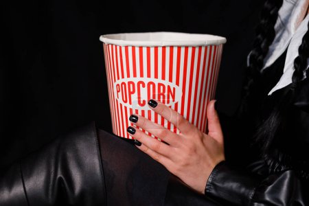 Foto de A brunette with two pigtails in a black dress holds a box of popcorn. Wednesday series from Netflix. Copy space - Imagen libre de derechos