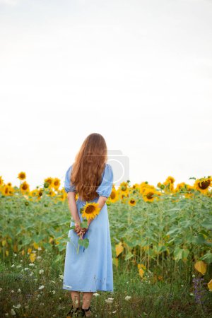 Foto de Vinnytsia, Ukraine - August 3, 2022. A girl in a blue dress on a sunflower field holds a flower in her hands - Imagen libre de derechos