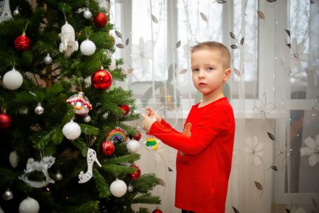 Foto de A boy decorates a Christmas tree with white and red toys. Waiting for a miracle and a holiday at home - Imagen libre de derechos