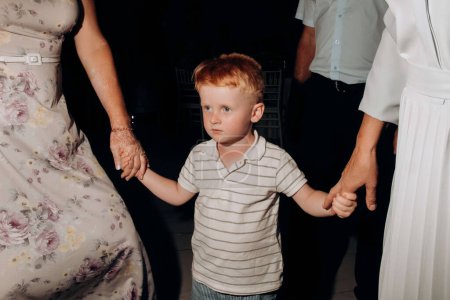 Foto de A red-haired boy holds the hands of two grandmothers while dancing at a wedding - Imagen libre de derechos