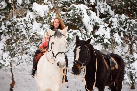 Foto de Red-haired girl on a horse in winter in the forest. Everything around is white. Christmas atmosphere around the world. Copy space - Imagen libre de derechos