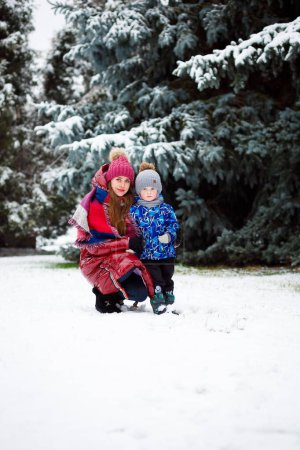 Foto de Una madre con una chaqueta roja abraza a su hijo en invierno en un bosque con mucha nieve. Año nuevo 2023 - Imagen libre de derechos