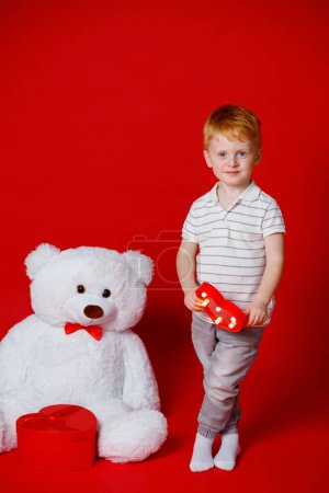 Foto de A red-haired 3-year-old boy holds a red heart in his hands for Valentine's Day. A child loves his parents, and they love him too - Imagen libre de derechos