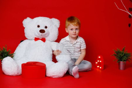 Foto de A red-haired boy with a white bear on a red background. Childhood in Ukraine. Russia started a war and all children are afraid of the sound of explosions - Imagen libre de derechos