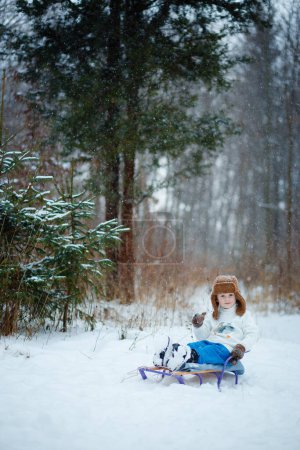 A 4-year-old boy sits on a sled in a snowy forest in winter. A warm brown hat, blue pants, a white fur collar