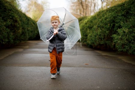 A red-haired boy walks with an umbrella in the rain in the park. Brown suit and gray jacket