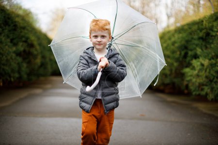 A red-haired boy walks with an umbrella in the rain in the park. Brown suit and gray jacket