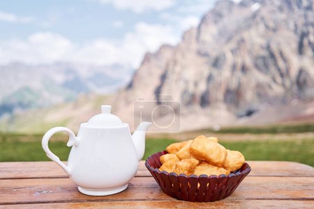 Téléchargez les photos : Théière avec thé et délicieuses pâtisseries kazakhes baursak. Café de rue dans les hauts plateaux avec vue sur les montagnes.. Faire frire la pâte dans de l'huile bouillante dans une poêle. Photo de haute qualité - en image libre de droit