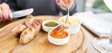 Photo for A piece of sausage is dipped in sauce. Lunch in a restaurant, a woman cuts Grilled sausages. Barbecue restaurant menu, a series of photos of different means. High quality photo - Royalty Free Image
