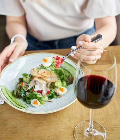 Photo for A woman is seated at a table with a plate of food and a glass of Burgundy wine in stemware - Royalty Free Image