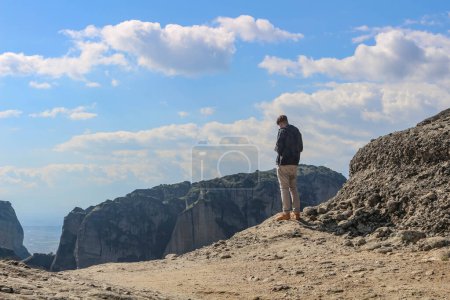 Experimente la emoción de la aventura mientras un turista se para en el borde, contemplando los impresionantes acantilados de Meteora, Grecia, en un día soleado y radiante.