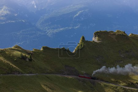 Téléchargez les photos : Amazing hiking day in the alps of Switzerland. A beautiful locomotive drives on to a mountain and a lot of smoke comes out of the locomotive. What a view in the background. - en image libre de droit