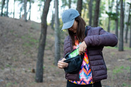young latina woman in jacket and cap holding or taking a cell phone out of a fanny pack in a forest