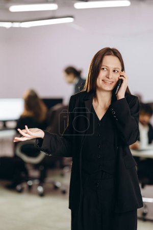 Foto de Female company ceo talking on the phone with smile. Beautiful businesswoman making phone call in office. Charming manager using smartphone at her workplace. High quality photo - Imagen libre de derechos