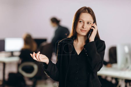 Téléchargez les photos : Femme entrepreneur parlant au téléphone sur fond flou. Belle femme d'affaires en tenue noire faisant un appel téléphonique dans son bureau. Photo de haute qualité - en image libre de droit