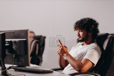 Photo for Office worker typing message on smartphone. Curly young man sitting at the computer desk in office and using his phone. High quality photo - Royalty Free Image