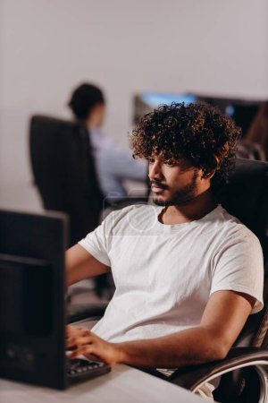 Foto de Focused curly guy working with computer in office. IT specialist typing on keyboard at his workplace. Young man in white t-shirt coding on blurred background. High quality photo - Imagen libre de derechos