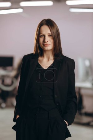 Photo for Confident businesswoman posing with hands in pockets. Indoor photo of successful office worker girl. Female manager in black suit on blurred background. High quality photo - Royalty Free Image