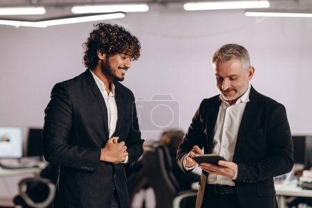 Photo for Smiling coworkers having small talk in office. Two businessmen in black suits with digital tablet. High quality photo - Royalty Free Image