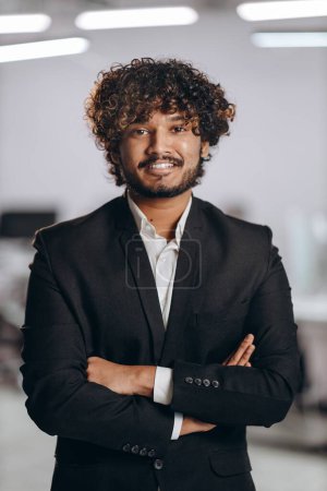 Téléchargez les photos : Cheerful young businessman posing with folded arms. Successful entrepreneur with curly hair looking at camera with smile. Happy Indian manager in black suit. High quality photo - en image libre de droit