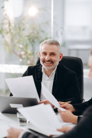 Photo for Smiling middle aged business man sitting in the office. A male businessman looks over documents and smiles during a business meeting in a conference room. High quality photo - Royalty Free Image