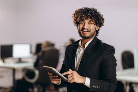 Photo for Excited businessman scrolls tablet screen. Indoor shot of smiling entrepreneur with digital device. Happy office worker looking at camera. High quality photo - Royalty Free Image