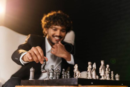 Téléchargez les photos : Vue d'un homme souriant en costume d'affaires jouant aux échecs. Un homme assis près d'un échiquier est sur le point de faire bouger un chevalier. Photo de haute qualité - en image libre de droit