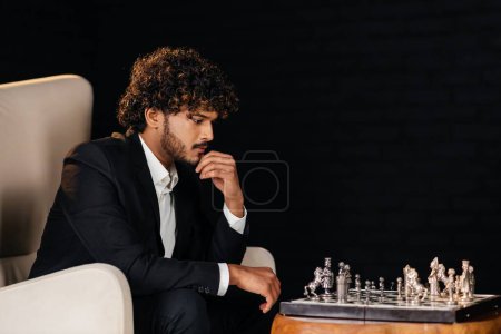 Téléchargez les photos : Un joueur d'échecs regarde un échiquier tout en envisageant des mouvements possibles. Un homme d'affaires concentré en tenue formelle s'assoit dans un fauteuil et joue aux échecs avec lui-même. Entrepreneur penser à - en image libre de droit