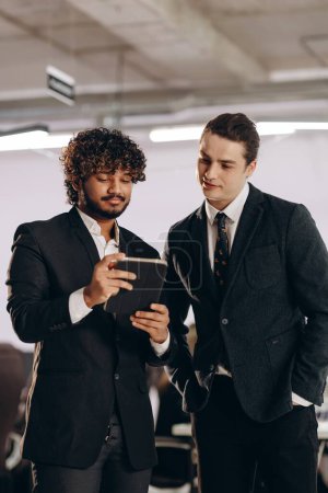 Foto de Curiosos hombres de negocios viendo vídeo en la tableta. Foto interior de compañeros de trabajo en trajes negros usando gadget digital en la oficina. Foto de alta calidad - Imagen libre de derechos