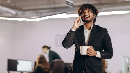 Foto de Un hombre de negocios sonriente con una taza de café hablando por teléfono. Gerente corporativo guapo usando smartphone en la oficina. Foto de alta calidad - Imagen libre de derechos