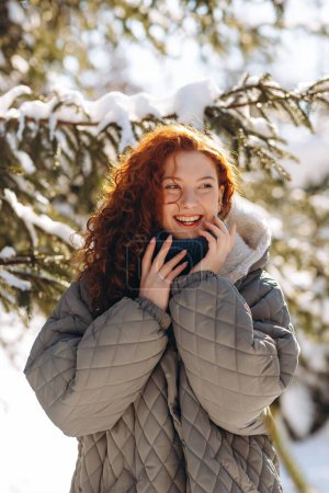 Téléchargez les photos : Vue d'une jolie femme souriante dans une veste en duvet gris. Une fille aux cheveux roux se promène dans le parc par une matinée ensoleillée d'hiver. Photo de haute qualité - en image libre de droit
