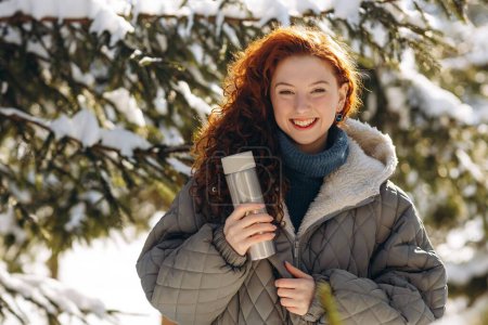 Photo for A smiling woman with a thermos in her hand stands against the backdrop of a snowy forest. Beautiful red-haired woman posing against the backdrop of snow-covered trees. High quality photo - Royalty Free Image