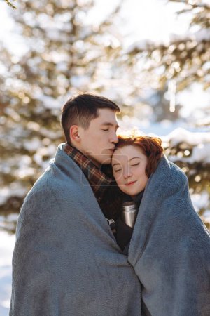 Téléchargez les photos : Les jeunes amoureux se tiennent au milieu d'un parc d'hiver ensoleillé enveloppé dans une couverture pour garder au chaud. Un jeune couple passe sa journée à marcher dans un parc enneigé. Photo de haute qualité - en image libre de droit