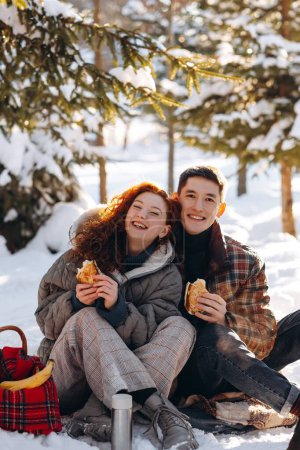 Téléchargez les photos : Heureux jeune couple ont un pique-nique romantique dans le parc d'hiver. Une jeune femme rousse boit du thé et mange des gâteaux faits maison avec son petit ami au milieu d'un parc enneigé. Photo de haute qualité - en image libre de droit