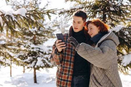 Foto de Un chico joven y una chica con ropa de invierno están mirando algo en un teléfono inteligente. El tipo y su novia están mirando fotos tomadas mientras caminan por un parque cubierto de nieve. Foto de alta calidad - Imagen libre de derechos