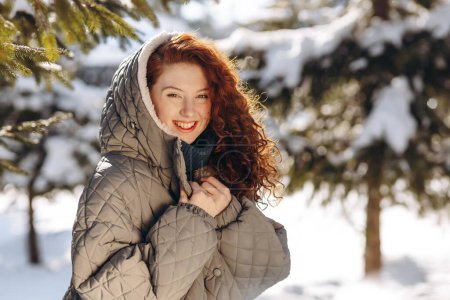 Photo for A cute red-haired woman in a hood poses against the backdrop of a winter park. Close-up of a young woman walking in the park on a winter sunny day. High quality photo - Royalty Free Image