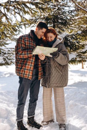 Téléchargez les photos : Deux jeunes hommes vêtus de vêtements d'hiver étudient une carte en se tenant au milieu d'une forêt enneigée. Perdu dans la forêt, un jeune couple étudie la carte pour trouver une sortie vers la route. Haut - en image libre de droit