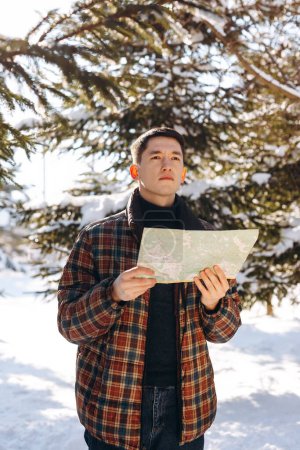 Téléchargez les photos : Un jeune homme vêtu d'une veste à carreaux se tient dans une forêt d'hiver avec une carte à la main. Un homme avec une carte dans les mains regarde quelque part au loin à la recherche de la bonne direction. Photo de haute qualité - en image libre de droit