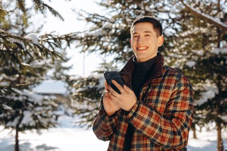 Photo for A smiling brunette man with a phone in his hands poses against the backdrop of a winter forest. View of a young man in a plaid down jacket standing in the middle of snow-covered trees. High quality - Royalty Free Image