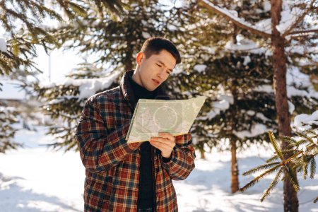 Photo for View of a young man standing in a snowy forest and looking at a map. A man lost in the winter forest is studying the map to find the nearest exit to the road. High quality photo - Royalty Free Image