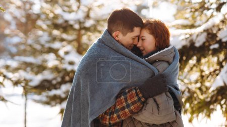 Téléchargez les photos : Photo romantique d'un jeune couple debout au milieu d'une forêt enneigée ensoleillée enveloppée dans une couverture. Un couple amoureux se tient sur le fond d'arbres enneigés, s'accrochant les uns aux autres - en image libre de droit