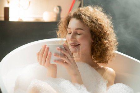 Photo for Smiling beautiful woman lies in a bath with foam and holds a cup with fragrant tea in her hands. A young woman with curly hair is taking a bubble bath and drinking chamomile tea. High quality photo - Royalty Free Image