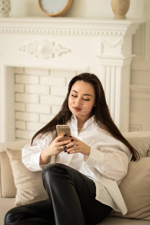 Foto de Una hermosa mujer de pelo largo se sienta en un sofá y navega por las redes sociales en un teléfono inteligente. Una mujer morena con una camisa blanca se comunica con sus amigos a través de un mensajero. Foto de alta calidad - Imagen libre de derechos