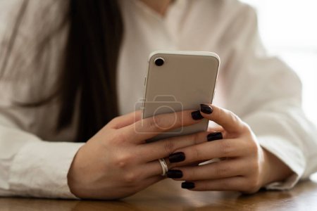 Photo for Cropped view of a woman in a white shirt who holds a white smartphone in her hands. A brunette woman sitting at a table uses a smartphone. High quality photo - Royalty Free Image