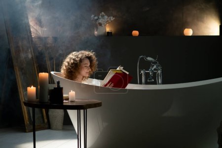 Photo for A young woman with curly hair takes a bath in a room with scented candles and reads a book. Beautiful woman is relaxing in the bath after a hard day at work. High quality photo - Royalty Free Image