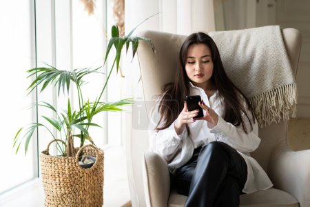 Photo for A young attractive brunette uses a smartphone while sitting in a chair near the window. A beautiful woman sits near a window with a plant and browses social networks on her smartphone. High quality - Royalty Free Image